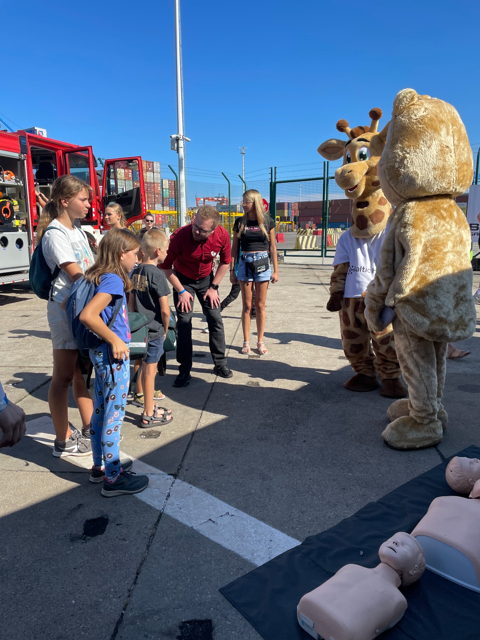 Instruktor w czerwonej koszuli demonstruje na manekinach zasady udzielania pierwszej pomocy grupie dzieci w Gdyni, obok stoi wóz strażacki i osoby przebrane za żyrafę i misia.