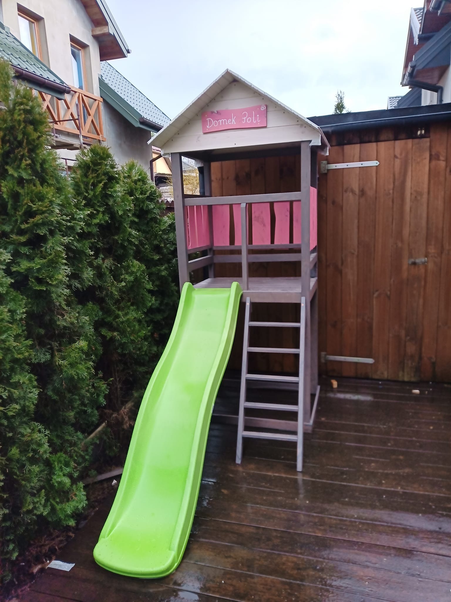 Drewniany plac zabaw dla dzieci z zieloną zjeżdżalnią i domkiem z napisem 'Domek Poli', ustawiony na tarasie z desek. Widoczne tuje i drewniane ogrodzenie.