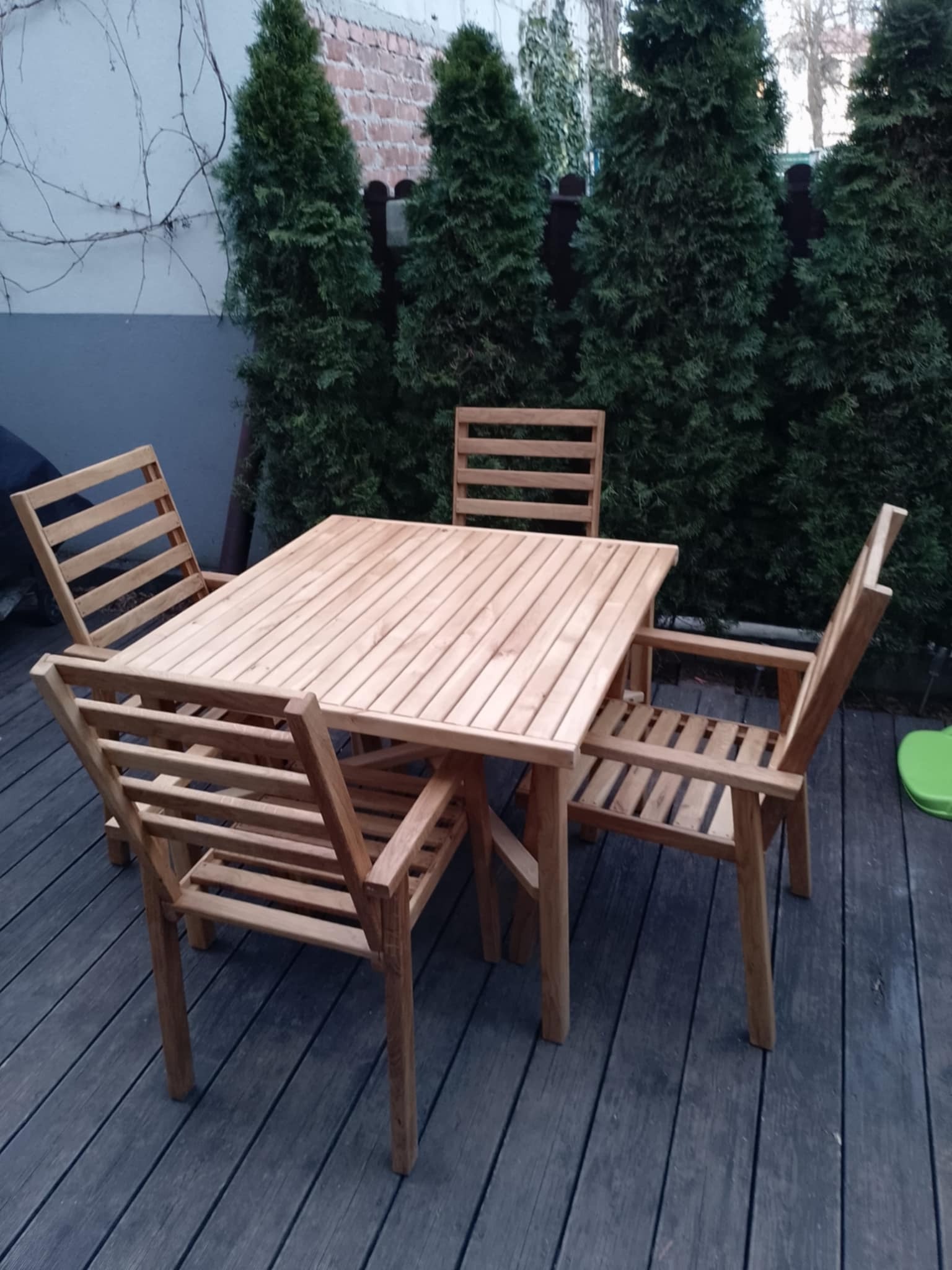 Drewniany stół i cztery krzesła z listew na tarasie z desek kompozytowych, w tle rząd wysokich tuj.