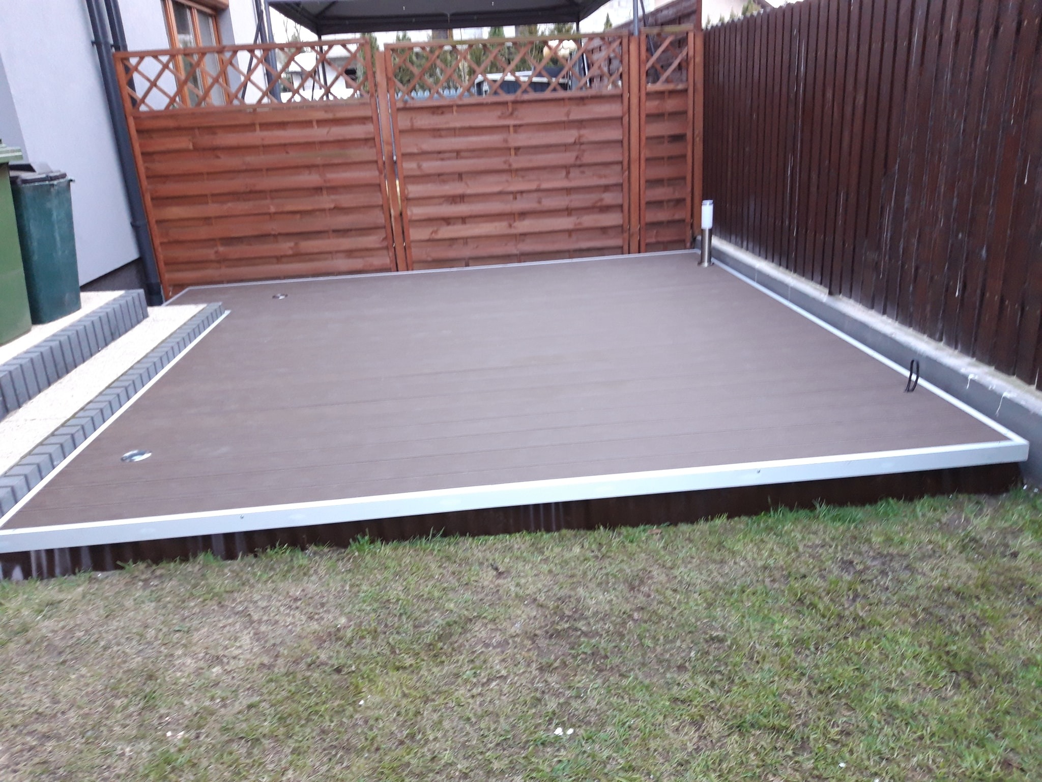 Nowoczesny taras z desek kompozytowych w kolorze brązowym, obrzeża z białego profilu aluminiowego, widok na drewniany płot i trawnik.