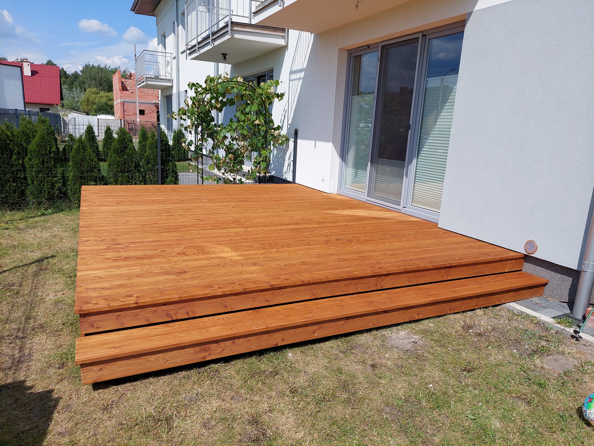 Nowoczesny taras z drewna o ciepłym, brązowym odcieniu, z dwoma stopniami, przylegający do białego budynku z przesuwnymi drzwiami tarasowymi, otoczony zielonym trawnikiem i żywopłotem.