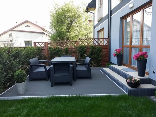 Nowoczesny taras z szarej deski kompozytowej, otoczony zielenią, z czarnymi meblami ogrodowymi i donicami z kwiatami przy wejściu do domu.