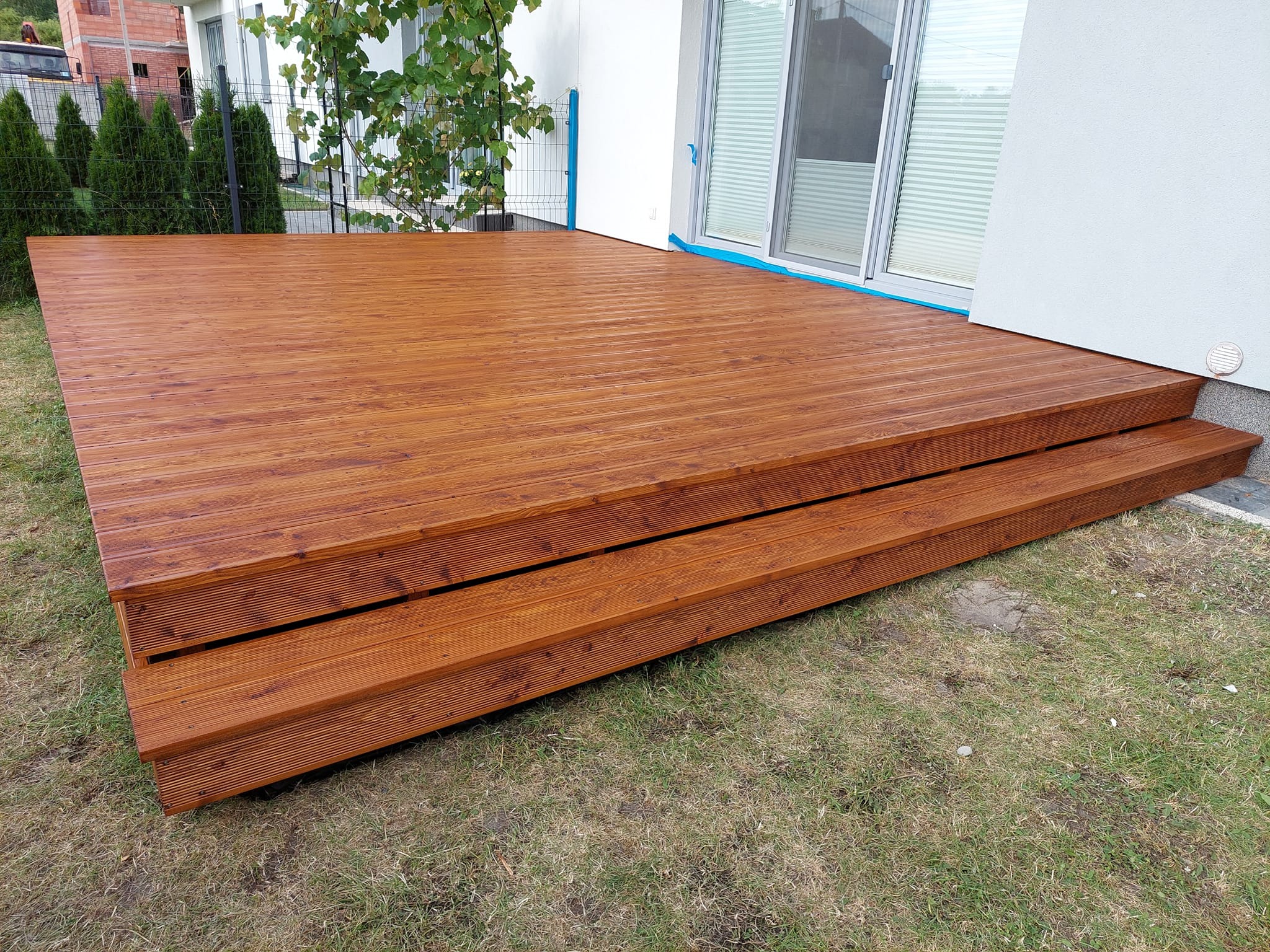 Nowy, brązowy taras z desek ryflowanych, z dwoma stopniami, przylegający do białego budynku z przesuwnymi drzwiami, otoczony zielenią i młodymi tujami.