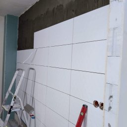Piotr Pawlak - Ściana łazienki w trakcie układania białych, poziomych płytek; widoczne fragmenty ściany z szarym klejem, drabina aluminiowa, poziomica i wystające rury instalacji wodnej.