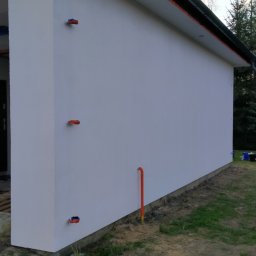 Piotr Pawlak - Boczny widok na otynkowaną ścianę budynku z widocznymi elementami instalacji elektrycznej i rurą wystającą z ziemi, otoczonego trawnikiem i drzewami.