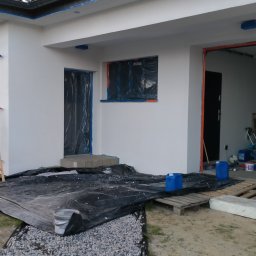 Piotr Pawlak - Biała elewacja domu jednorodzinnego w trakcie prac przygotowawczych, okna i drzwi zabezpieczone folią, teren wokół przykryty czarną plandeką i wysypany żwirem, widoczne niebieskie kanistry i paleta.