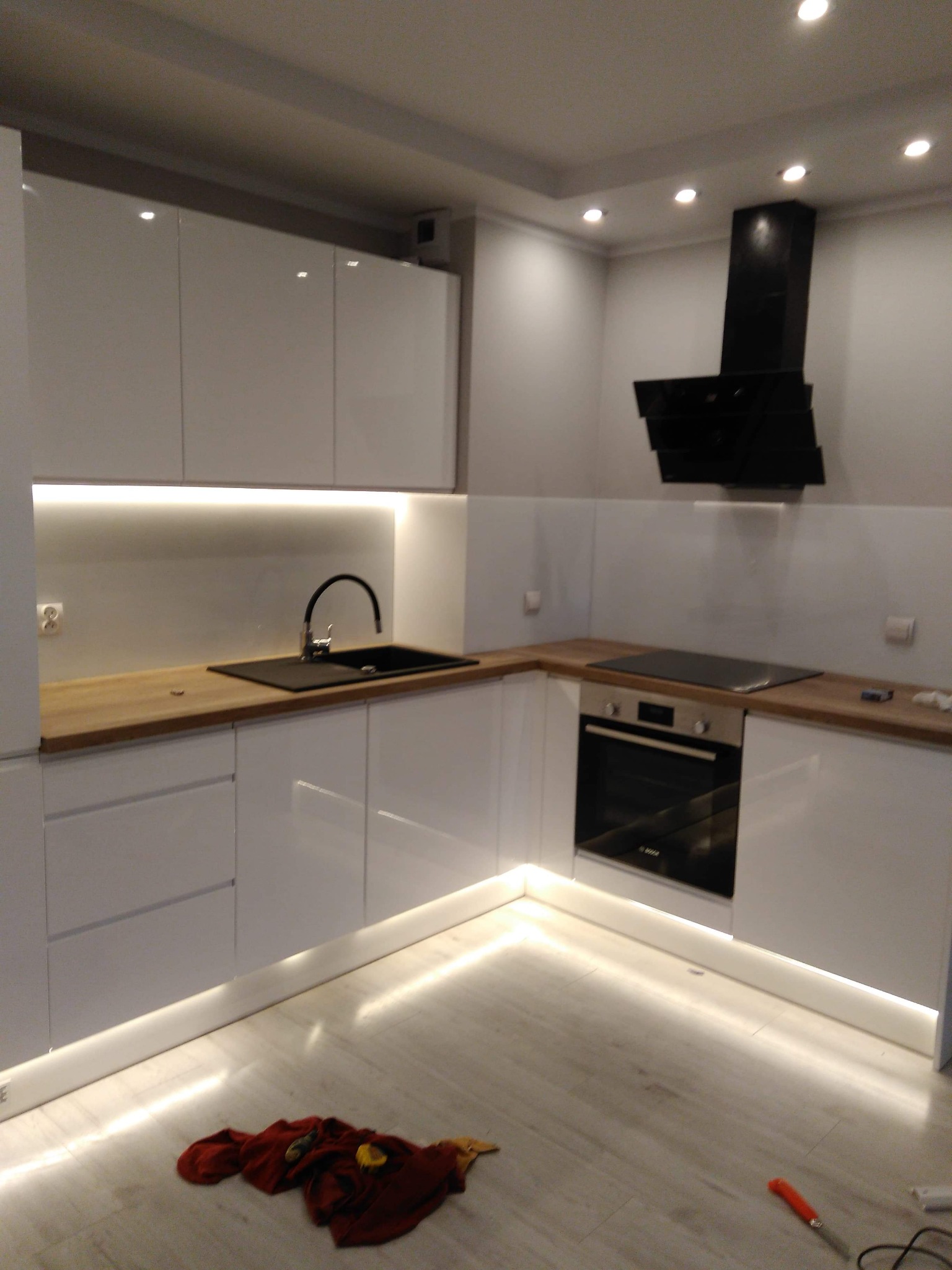 Nowoczesna kuchnia z białymi, lakierowanymi szafkami, drewnianym blatem, czarnym zlewem i płytą indukcyjną; oświetlenie LED pod szafkami i przy podłodze, widoczne narzędzie na podłodze.