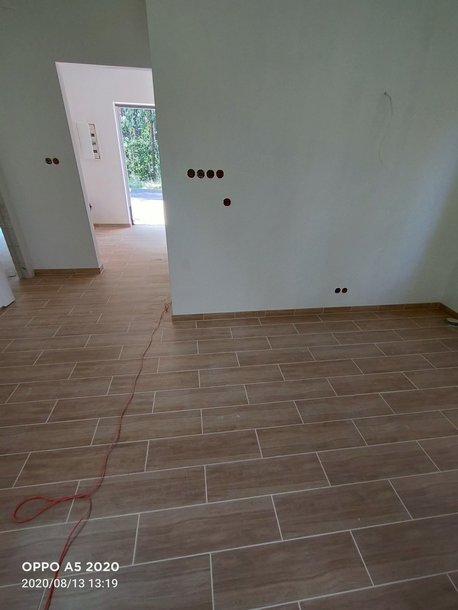 Wnętrze w trakcie wykańczania: brązowe płytki podłogowe imitujące drewno, białe ściany z widocznymi otworami na gniazdka elektryczne, czerwony kabel leżący na podłodze, widok na kolejne...