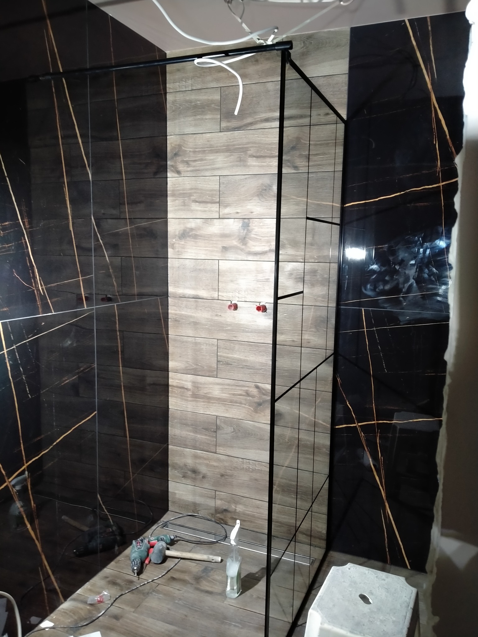 Częściowo wykończona kabina prysznicowa z czarnymi płytkami imitującymi marmur z złotymi żyłami oraz płytkami drewnopodobnymi, widoczne narzędzia i akcesoria glazurnicze na podłodze, szklane drzwi...