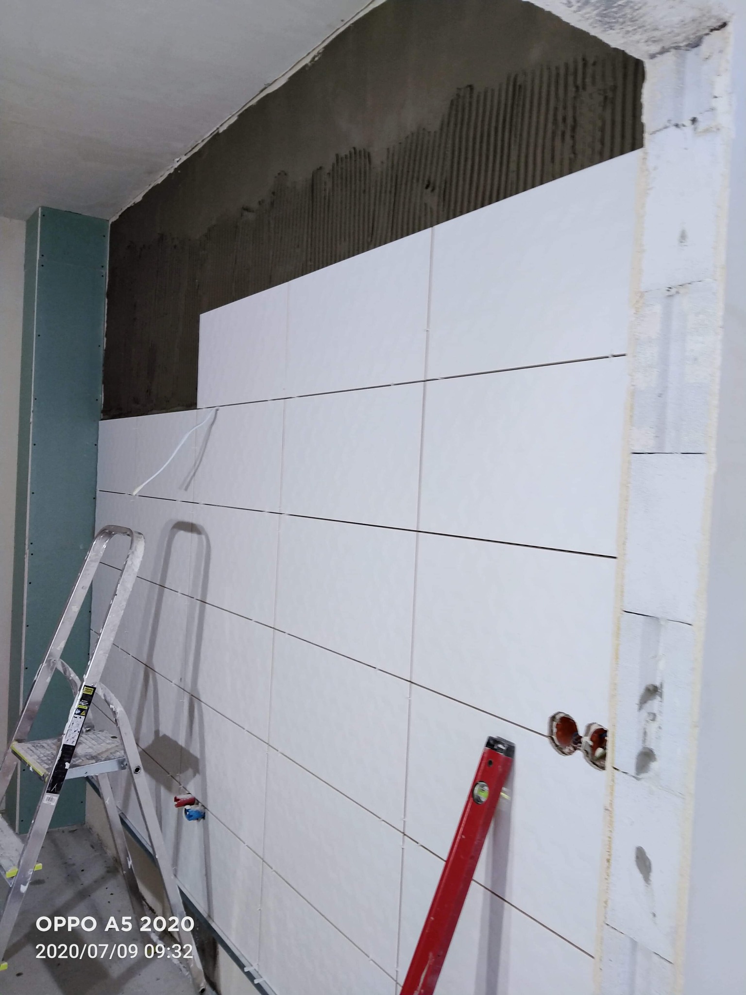 Ściana łazienki w trakcie układania białych, poziomych płytek; widoczne fragmenty ściany z szarym klejem, drabina aluminiowa, poziomica i wystające rury instalacji wodnej.
