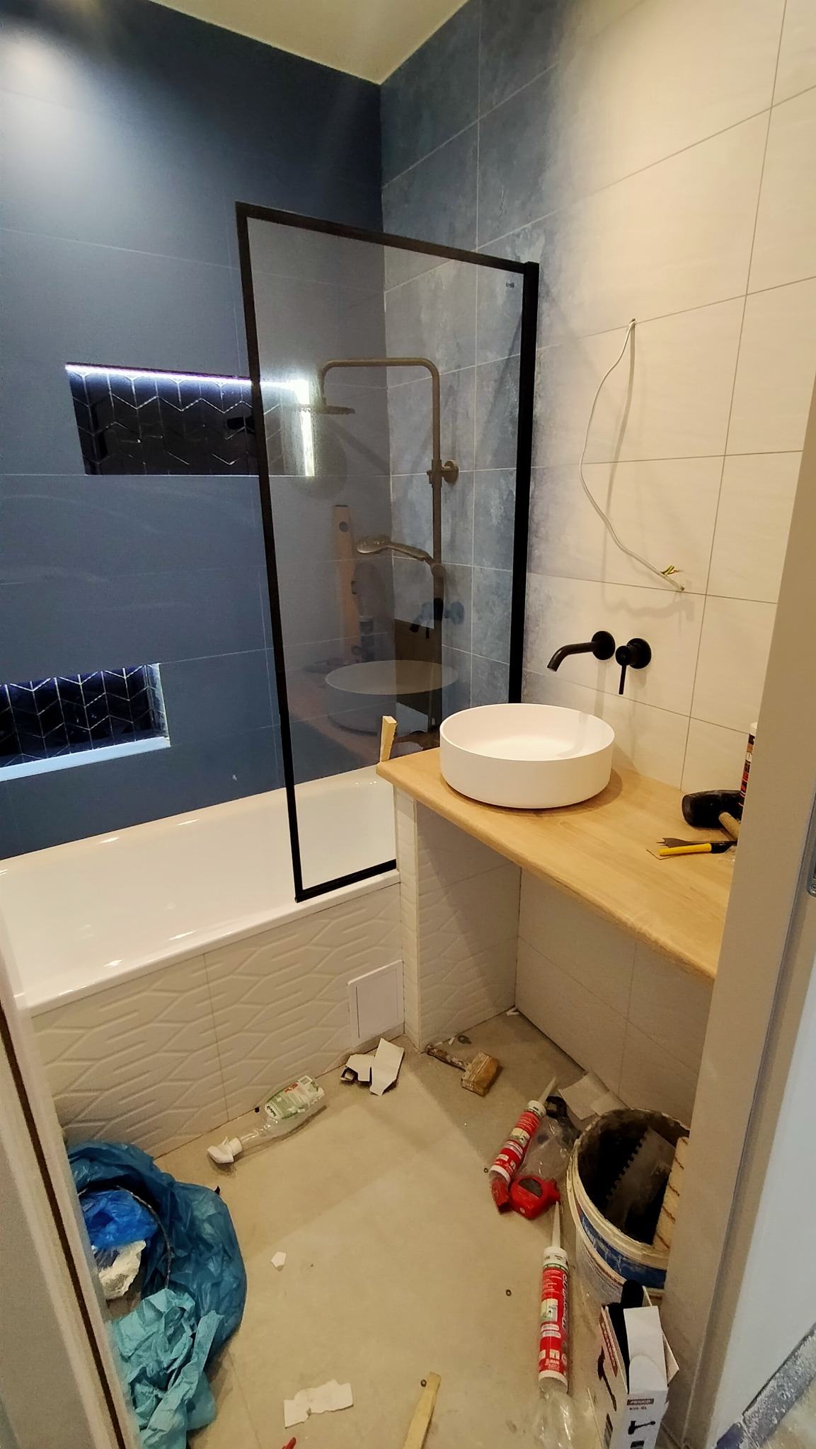 Nowoczesna łazienka w trakcie wykańczania: kabina prysznicowa z czarną ramą, umywalka nablatowa na drewnianym blacie, niebieskie i białe płytki na ścianach, widoczne narzędzia i materiały budowlane...