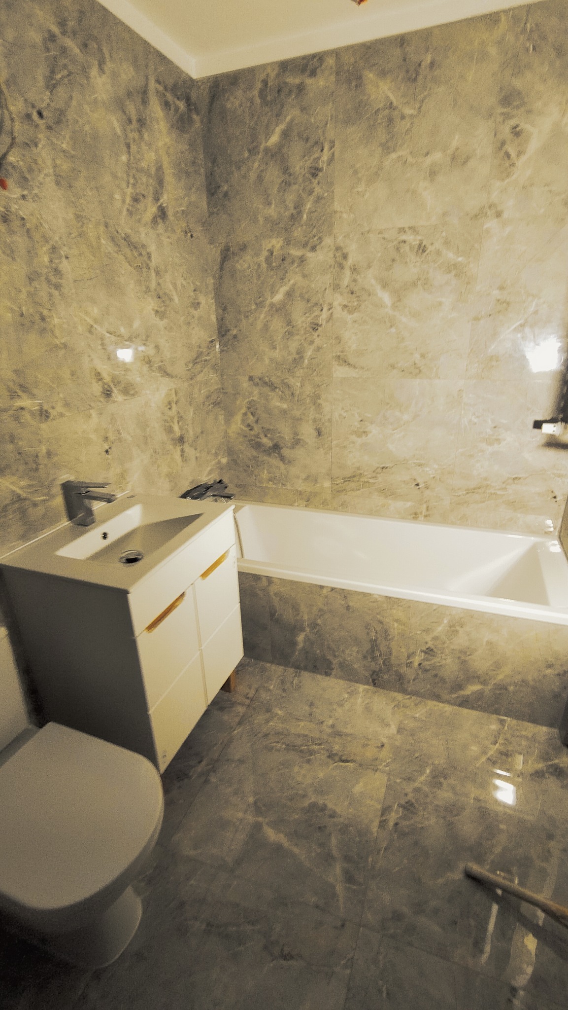 Wykończona łazienka w szarym marmurze z białą wanną, umywalką z szafką i toaletą; widoczne narzędzia glazurnicze na umywalce.