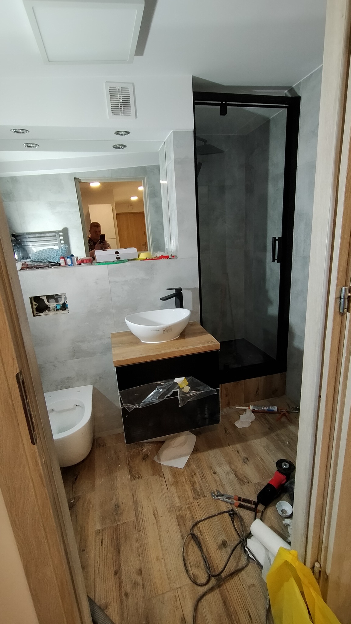Łazienka w trakcie prac: biała umywalka na drewnianym blacie z czarną szafką, kabina prysznicowa z czarną ramą, widoczne narzędzia i materiały budowlane na podłodze imitującej drewno, odbicie osoby...