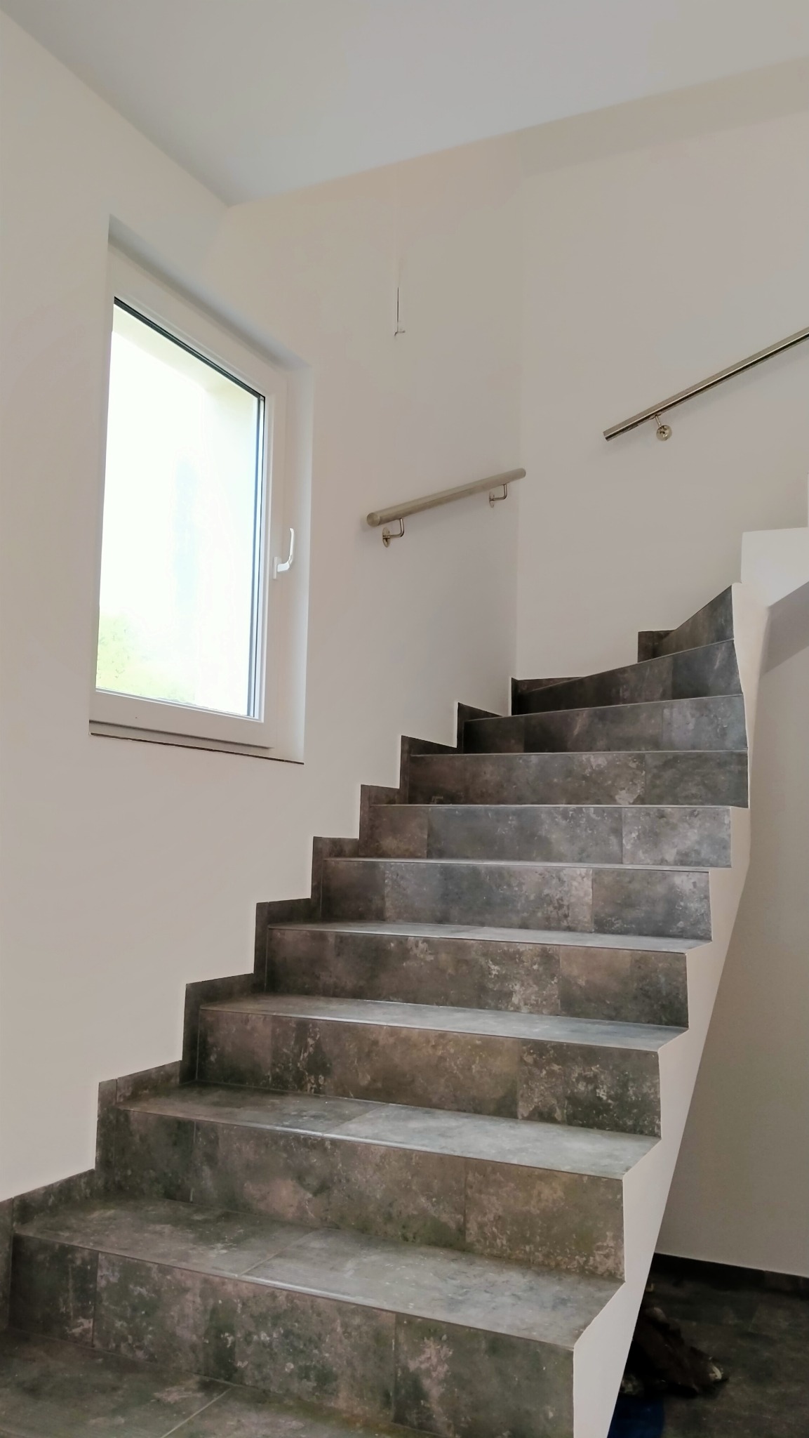 Klatka schodowa wyłożona szarymi płytkami imitującymi kamień, z metalową poręczą, oświetlona przez okno z białymi ramami, widok z dołu.