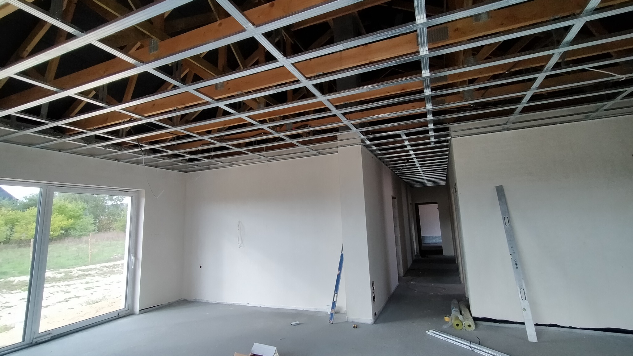 Wnętrze w trakcie prac wykończeniowych; widoczna metalowa konstrukcja sufitu podwieszanego, gładkie, białe ściany, duże okno balkonowe z widokiem na zieleń oraz pionowa poziomica oparta o ścianę.