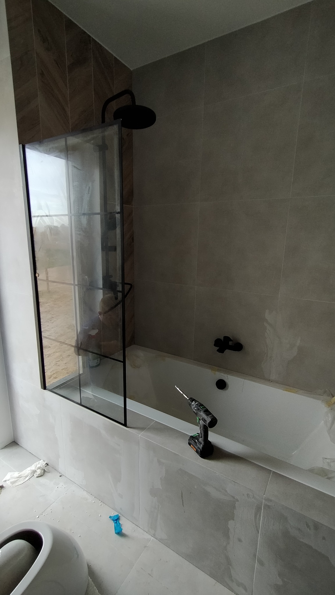 Wanna z zamontowaną czarną kabiną prysznicową typu walk-in, czarna deszczownica i bateria, na brzegu wanny stoi wiertarko-wkrętarka; widoczne ślady po fugowaniu płytek.