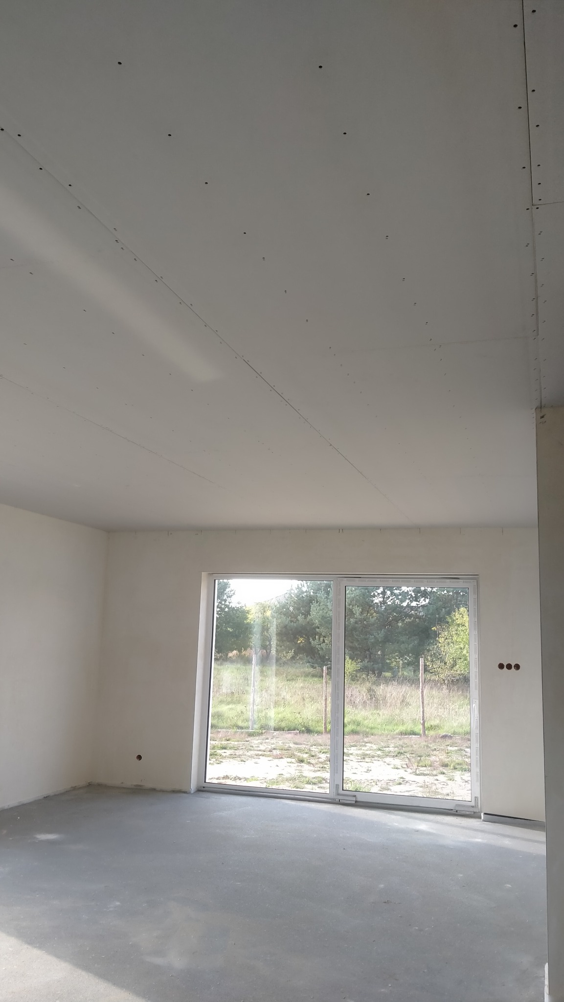 Surowe wnętrze z zamontowaną płytą karton-gips na suficie, widoczne łączenia i śruby, duże okno przesuwne z widokiem na zewnątrz.