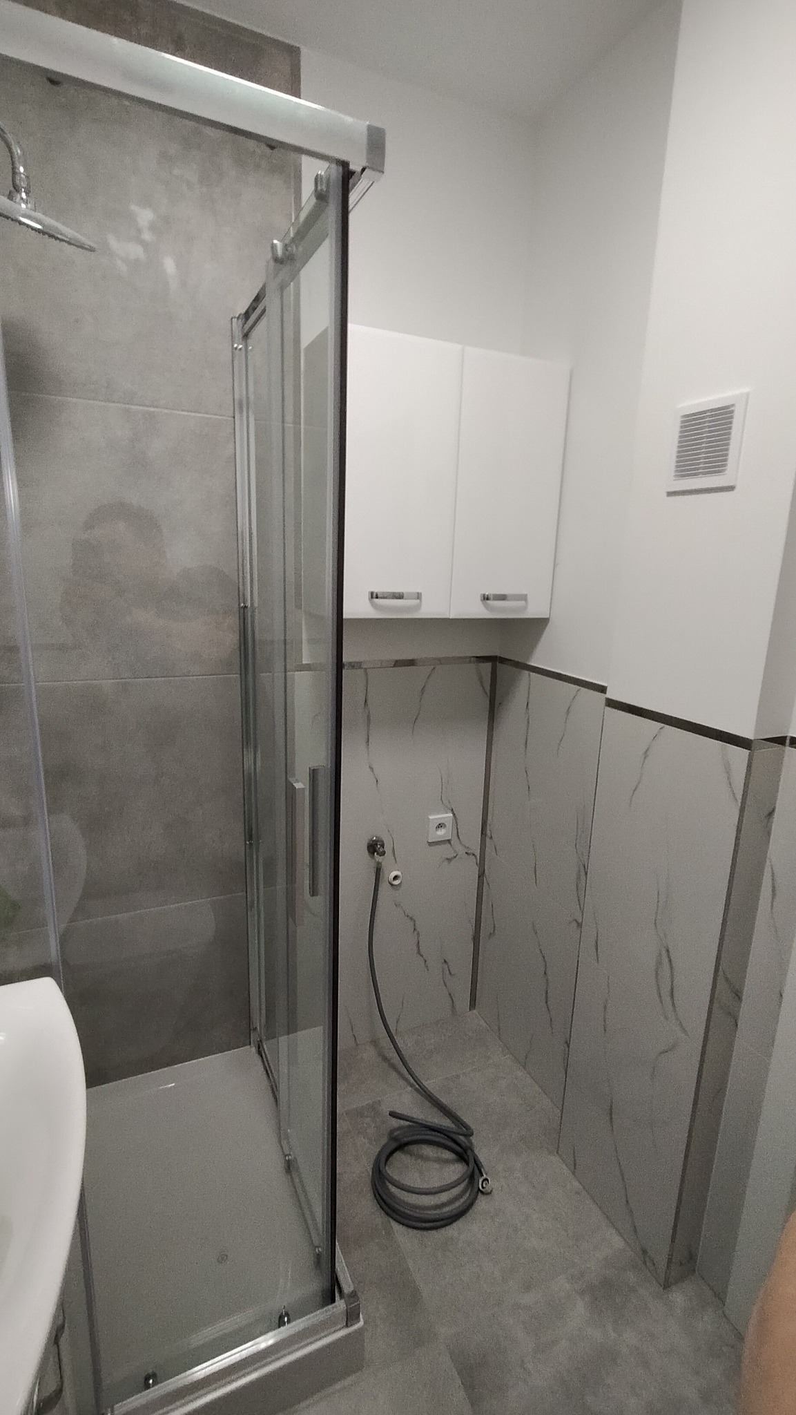 Szara łazienka z kabiną prysznicową z przesuwnymi drzwiami, białe szafki wiszące, płytki imitujące marmur na ścianach, wąż prysznicowy zwinięty na podłodze.
