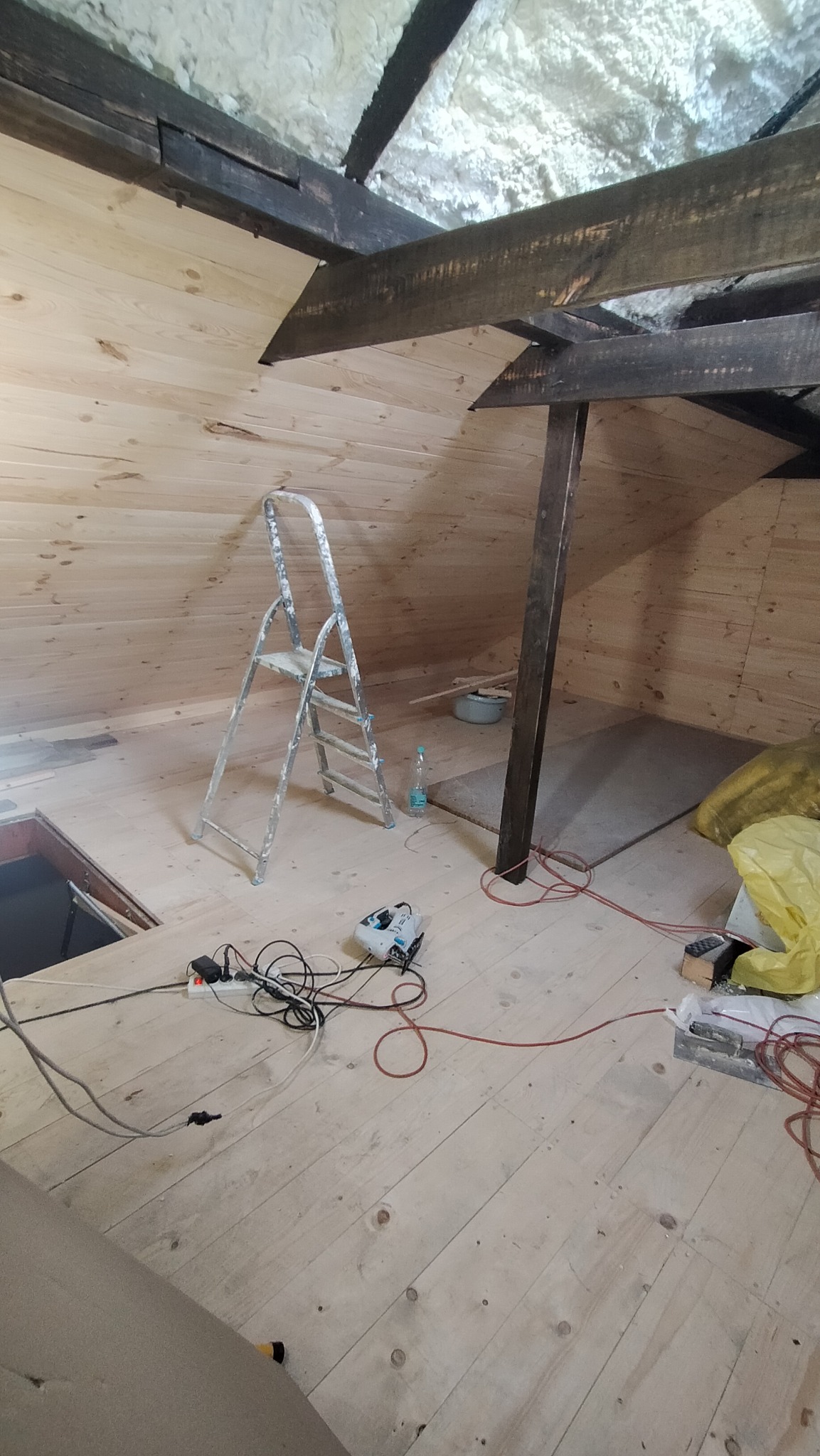Adaptacja poddasza: drewniana boazeria na ścianach i podłodze, aluminiowa drabina, widoczne krokwie, izolacja piankowa na suficie, narzędzia i kable na podłodze.