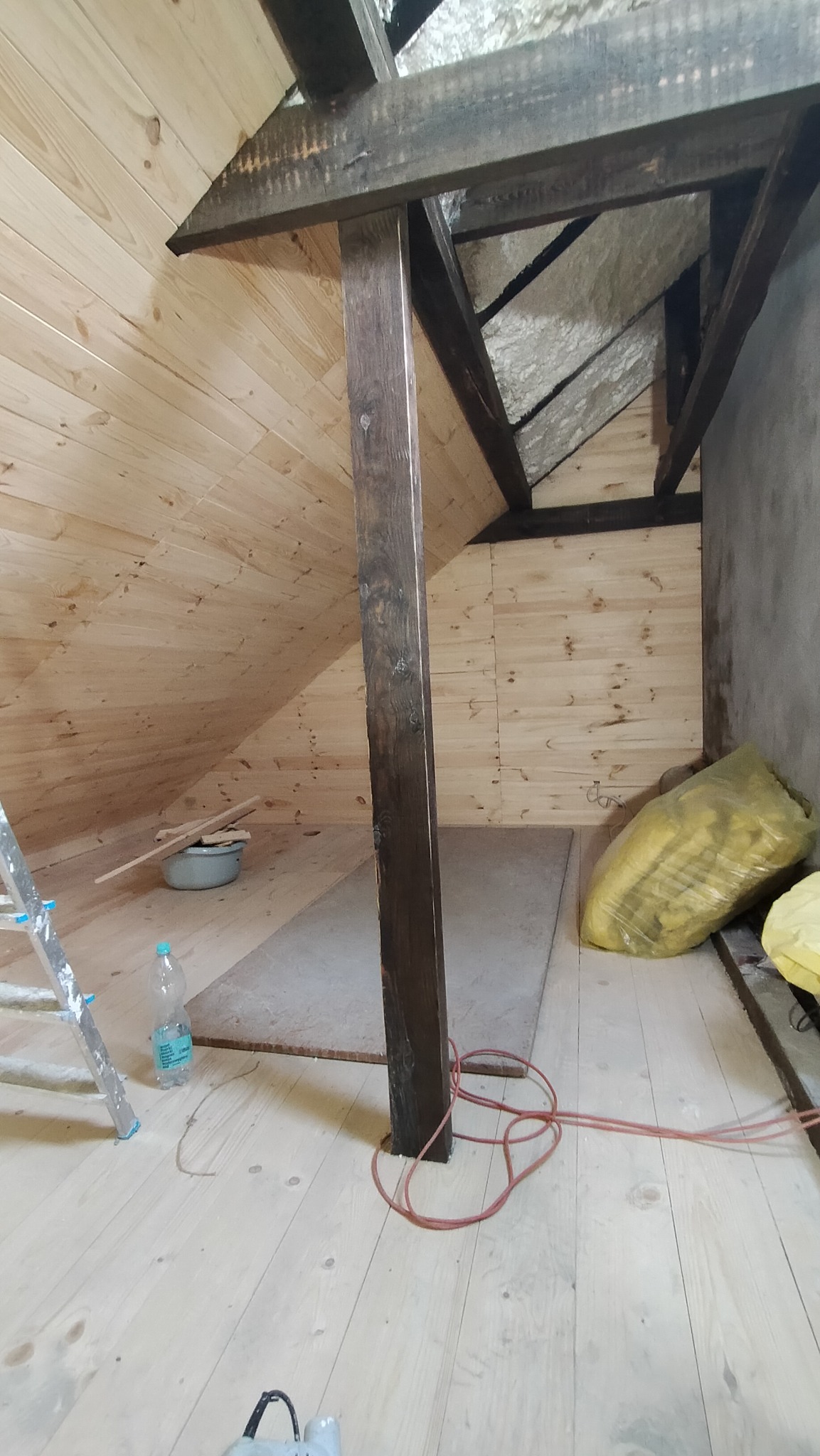 Adaptacja poddasza: drewniana boazeria na skośnej ścianie i suficie, widoczne krokwie, ocieplenie, kabel elektryczny na podłodze, drabina malarska i butelka wody mineralnej.