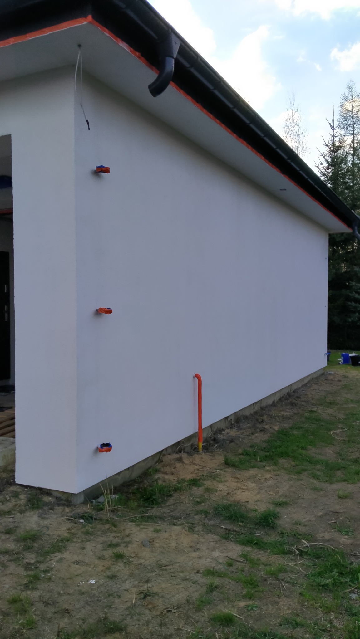 Boczny widok na otynkowaną ścianę budynku z widocznymi elementami instalacji elektrycznej i rurą wystającą z ziemi, otoczonego trawnikiem i drzewami.