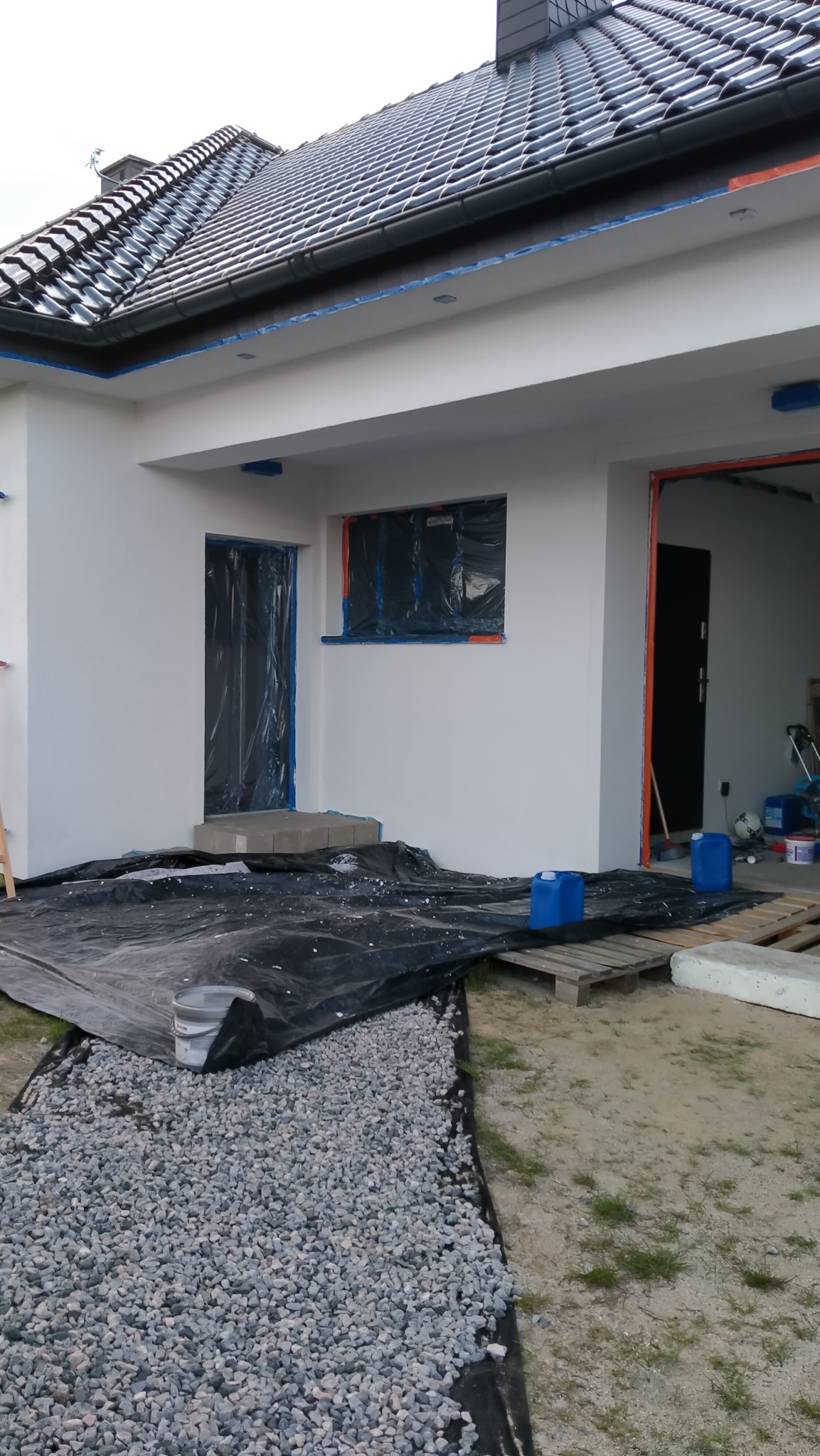 Biała elewacja domu jednorodzinnego w trakcie prac przygotowawczych, okna i drzwi zabezpieczone folią, teren wokół przykryty czarną plandeką i wysypany żwirem, widoczne niebieskie kanistry i paleta.