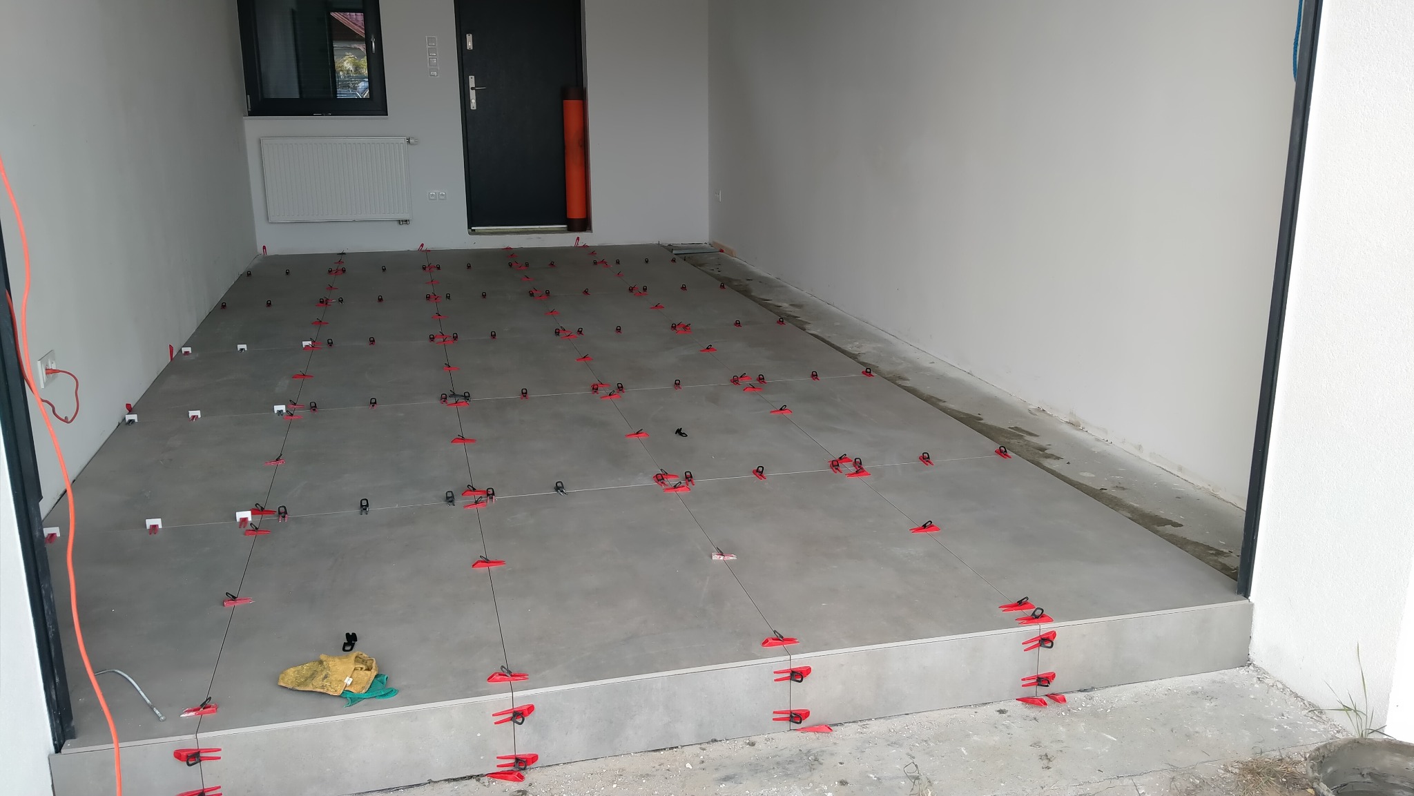 Szare płytki podłogowe w trakcie układania, z użyciem czerwonych klipsów poziomujących, widoczne narzędzia glazurnicze i kabel elektryczny przy ścianie, szara ściana i czarne drzwi w tle.