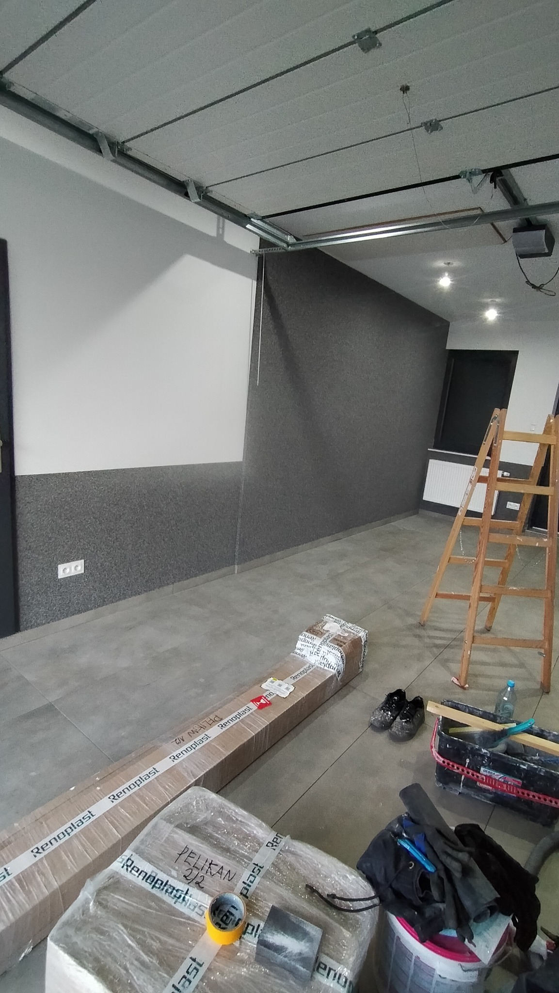 Wnętrze w trakcie wykończenia: częściowo otynkowana ściana na szaro, biała ściana powyżej, kartony z materiałami budowlanymi Renoplast, drabina malarska i narzędzia na szarych płytkach podłogowych.