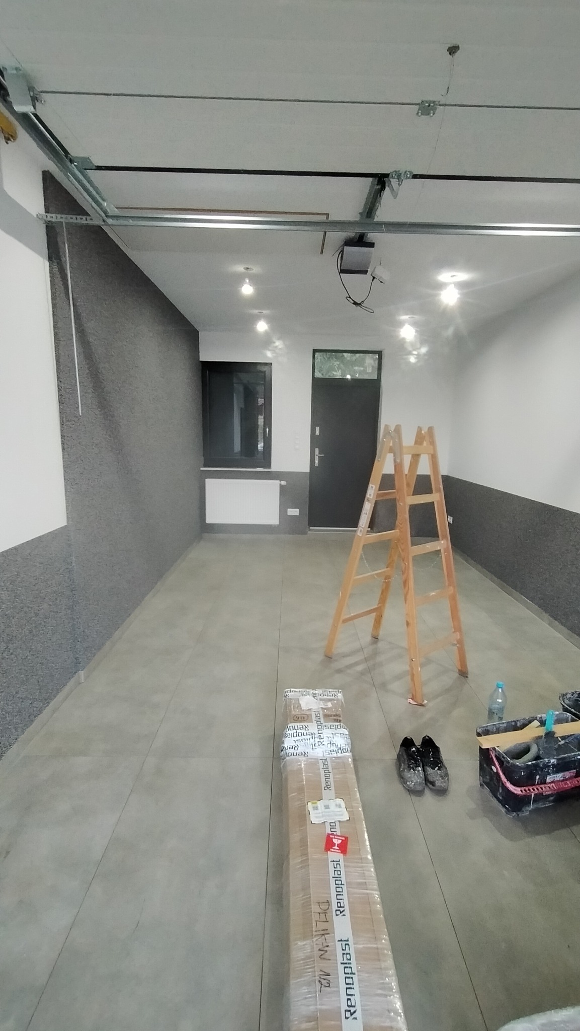 Wnętrze w Międzyrzeczu w trakcie prac wykończeniowych: częściowo położona struktura na ścianie, drabina, paczka paneli podłogowych Renoplast, para brudnych butów roboczych i wiadro z narzędziami...