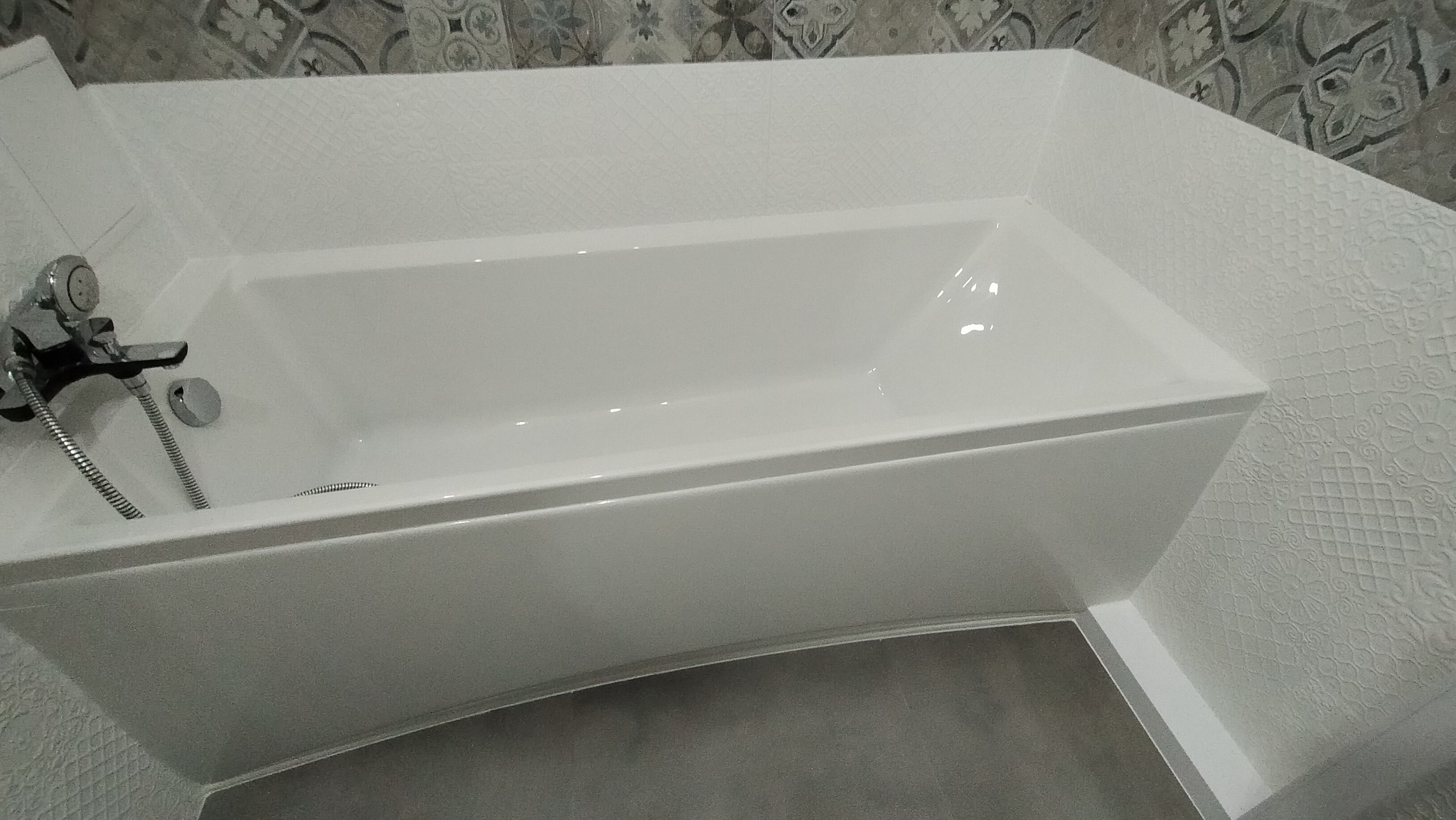 Nowoczesna, biała wanna z chromowaną baterią i szarą podłogą, otoczona ścianami wyłożonymi płytkami o zróżnicowanych wzorach i fakturach.