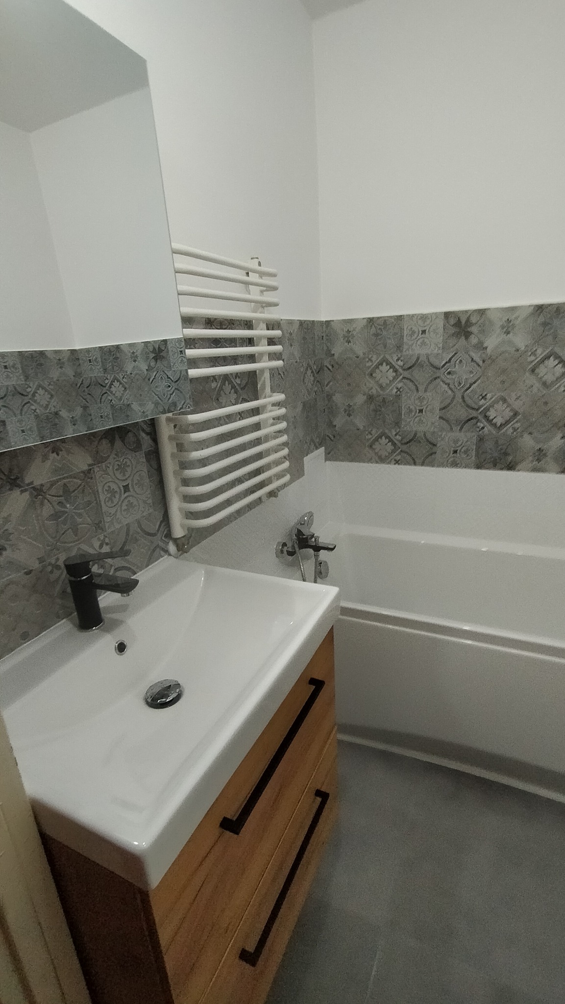 Nowoczesna łazienka po remoncie: biała umywalka z czarną baterią na szafce z drewnianymi szufladami, grzejnik drabinkowy na ścianie z szaro-białymi płytkami wzorzystymi, biała wanna.