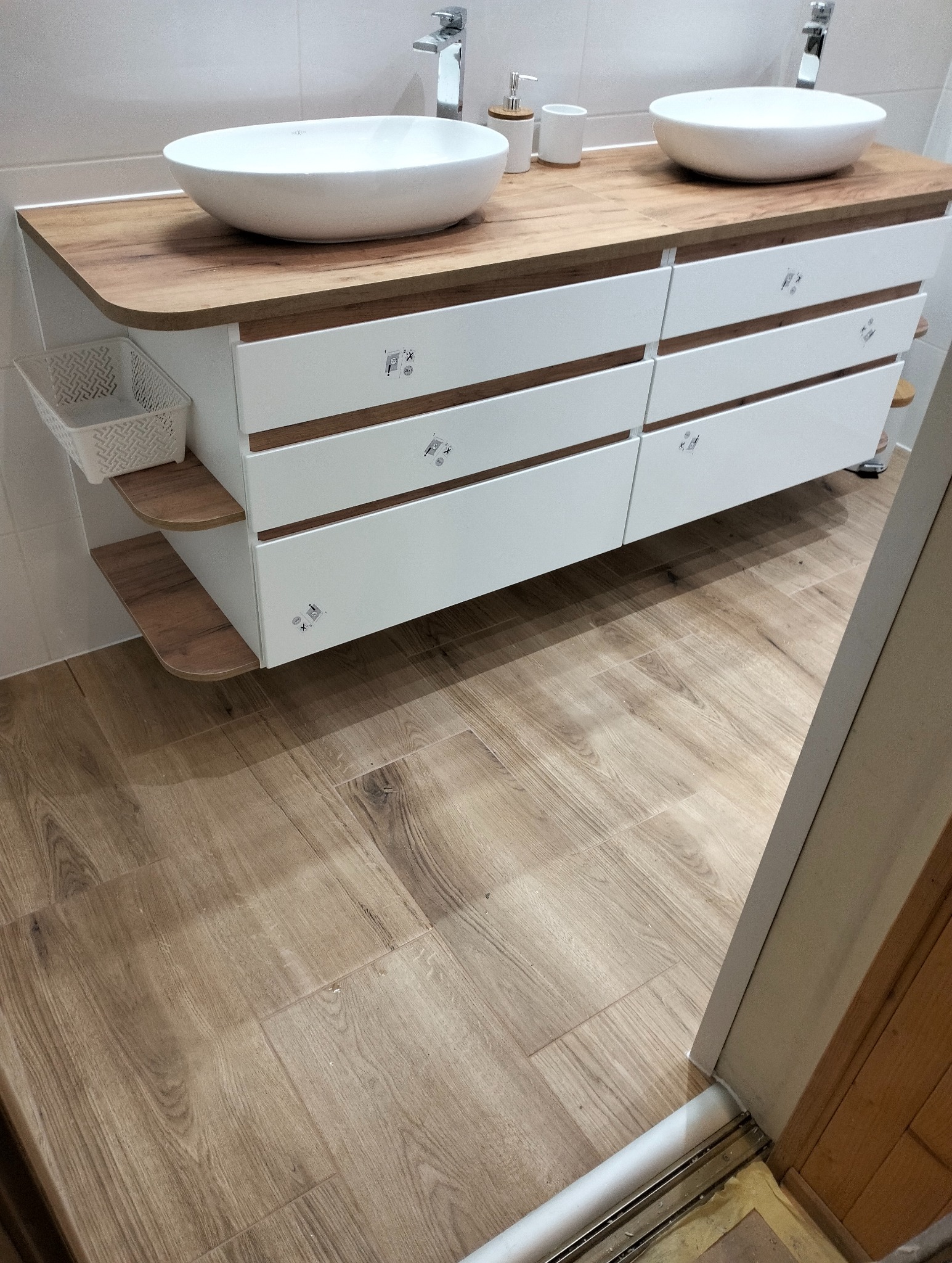 Nowoczesna łazienka z drewnianą podłogą i szafką pod umywalki nablatowe z sześcioma białymi szufladami, dodatkowymi półkami i koszykiem z boku.