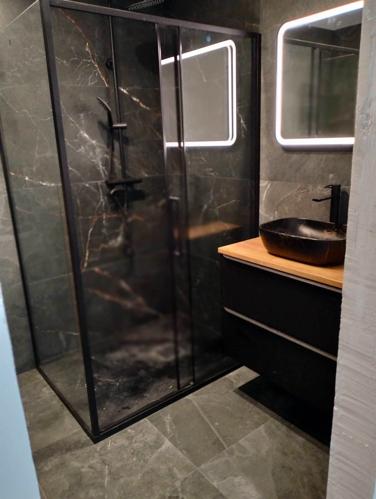 Nowoczesna łazienka z kabiną prysznicową typu walk-in z czarnymi ramami, marmurowymi płytkami na ścianach i podłodze, czarną umywalką na drewnianym blacie i podświetlanym lustrem.