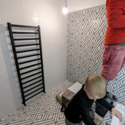 Czarny grzejnik łazienkowy na tle białych płytek i geometrycznego wzoru. Dziecko przygląda się pracy dorosłego na drabinie w trakcie montażu umywalki.