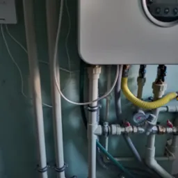 Szczegół instalacji grzewczej: biały piec gazowy z widocznymi rurami, zaworami i manometrem na tle zielonej ściany. Żółty przewód gazowy dodaje charakteru.