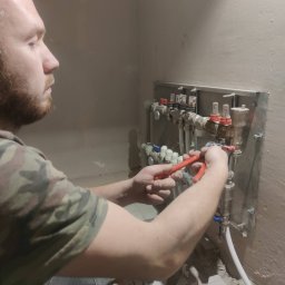 Sławomir Czarnulicz - Hydraulik z brodą, w koszulce moro, montuje rozdzielacz ogrzewania podłogowego, używając czerwonych szczypiec. Widoczne białe rury i surowa ściana.