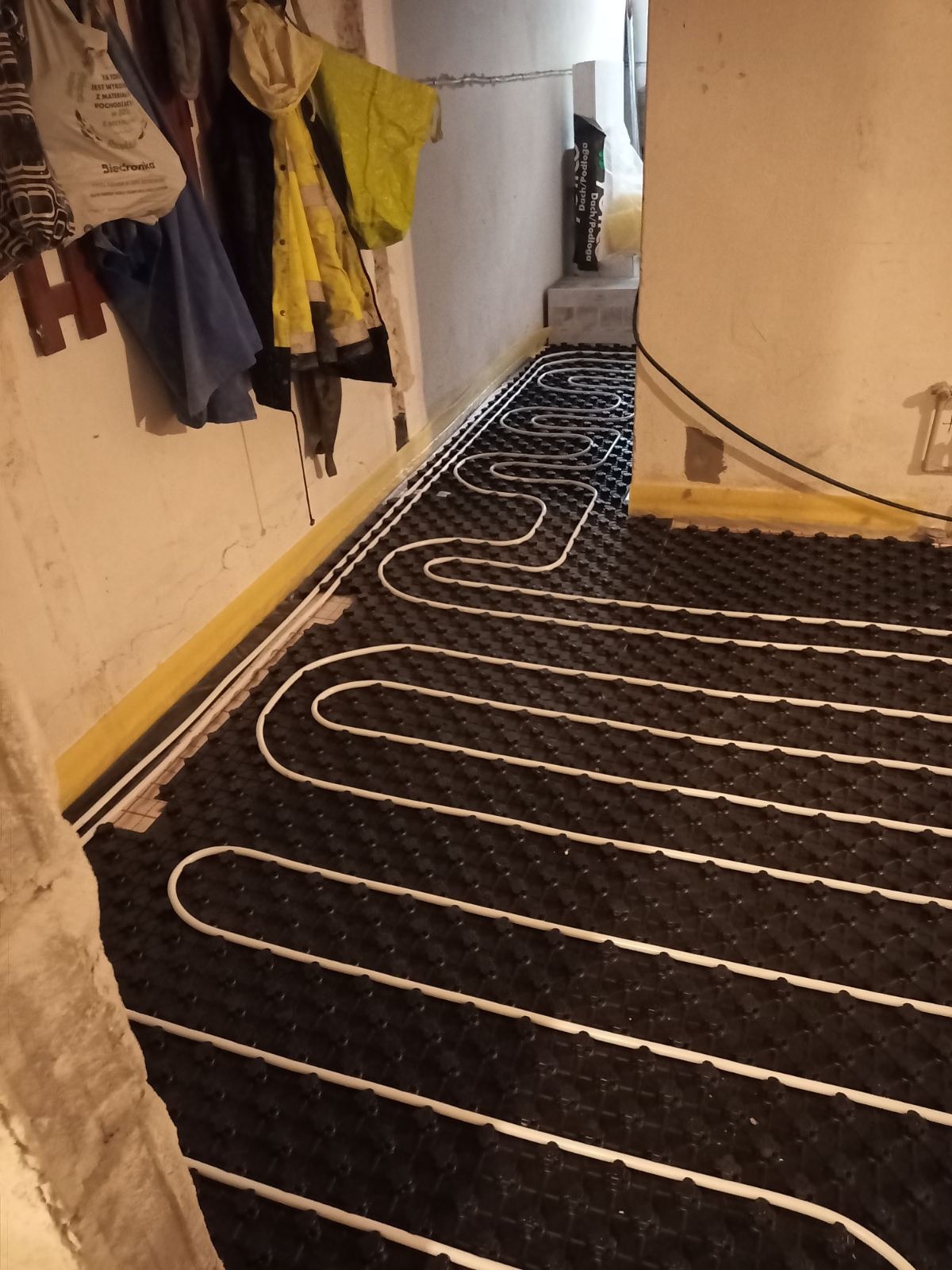 Białe rury ogrzewania podłogowego ułożone na czarnej macie izolacyjnej wzdłuż żółtej ściany. Widok z góry, wnętrze w trakcie prac instalacyjnych.