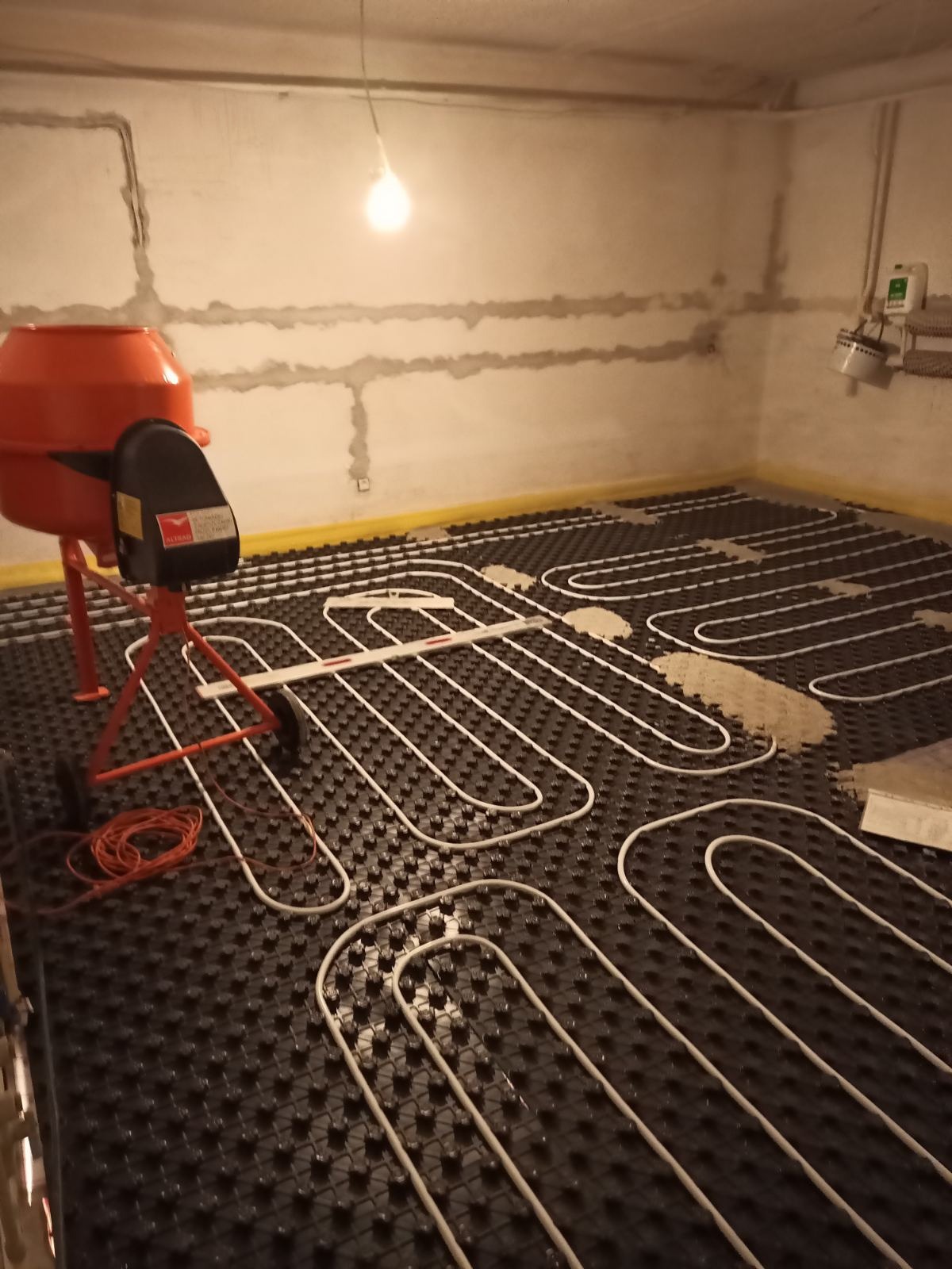 Instalacja ogrzewania podłogowego w pomieszczeniu z betoniarką. Rury ułożone na czarnej macie, częściowo zalane wylewką. Widoczne ściany w stanie surowym.