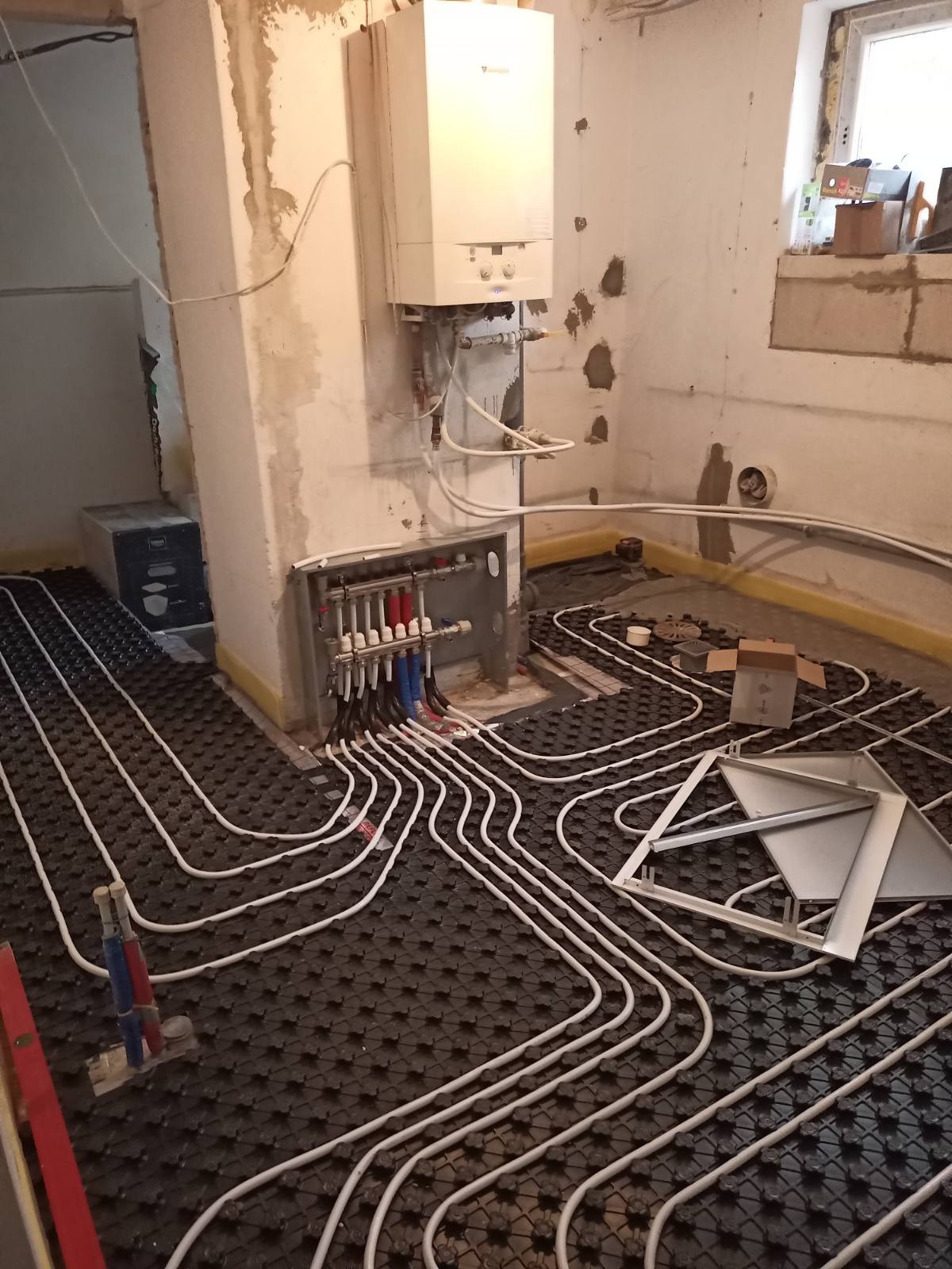 Białe rury ogrzewania podłogowego ułożone na czarnej macie, z rozdzielaczem i piecem gazowym w tle, w surowym wnętrzu z oknem.