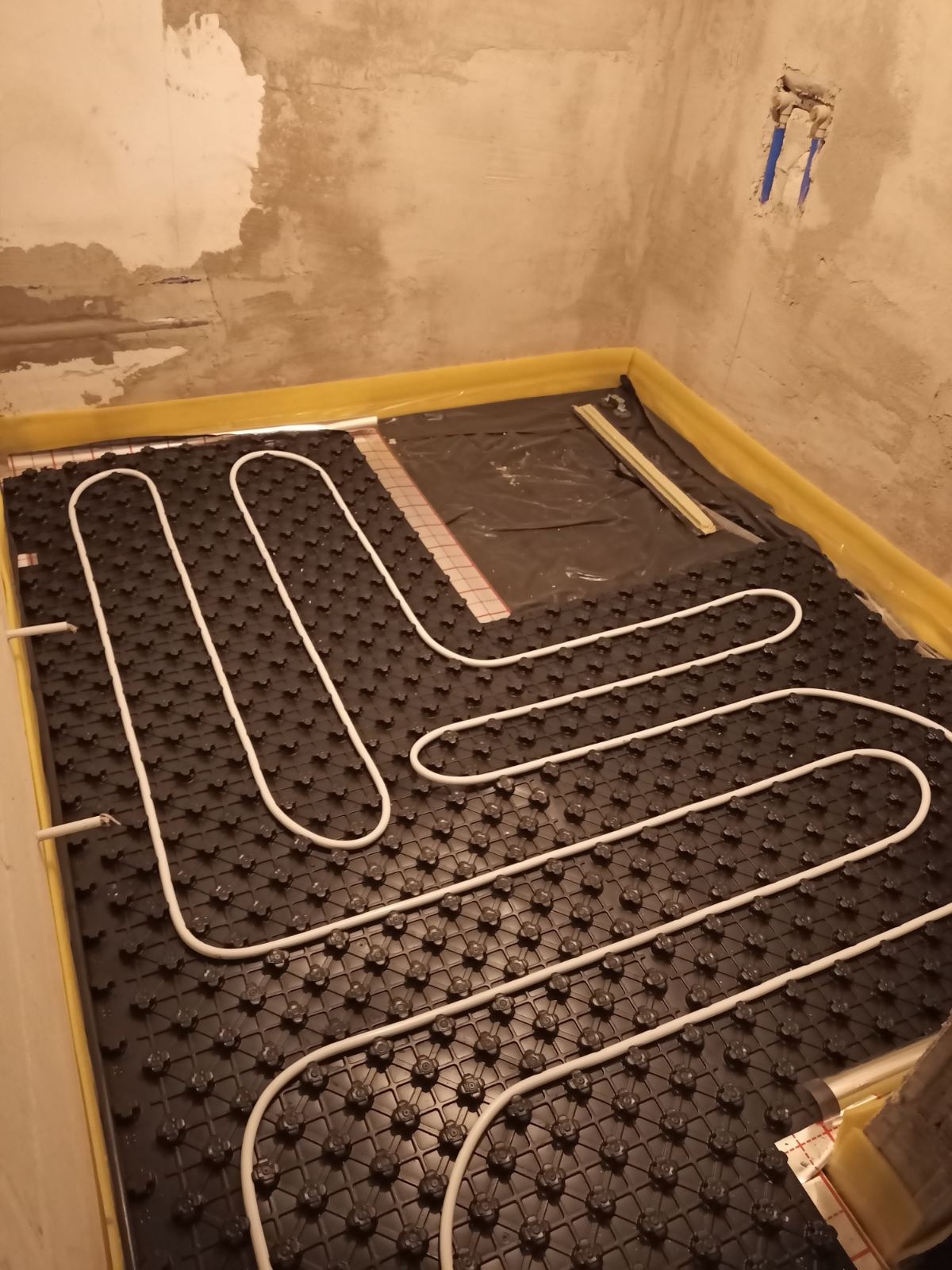 Białe rury ogrzewania podłogowego ułożone na czarnej macie z żółtą taśmą brzegową, widok z góry, surowa ściana w tle z widoczną instalacją.