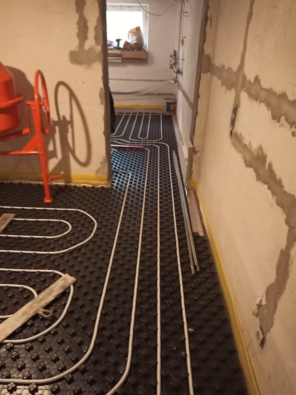 Instalacja ogrzewania podłogowego z widoczną pomarańczową betoniarką. Rury ułożone na czarnej macie z wypustkami, ściany surowe, przygotowane do tynkowania.