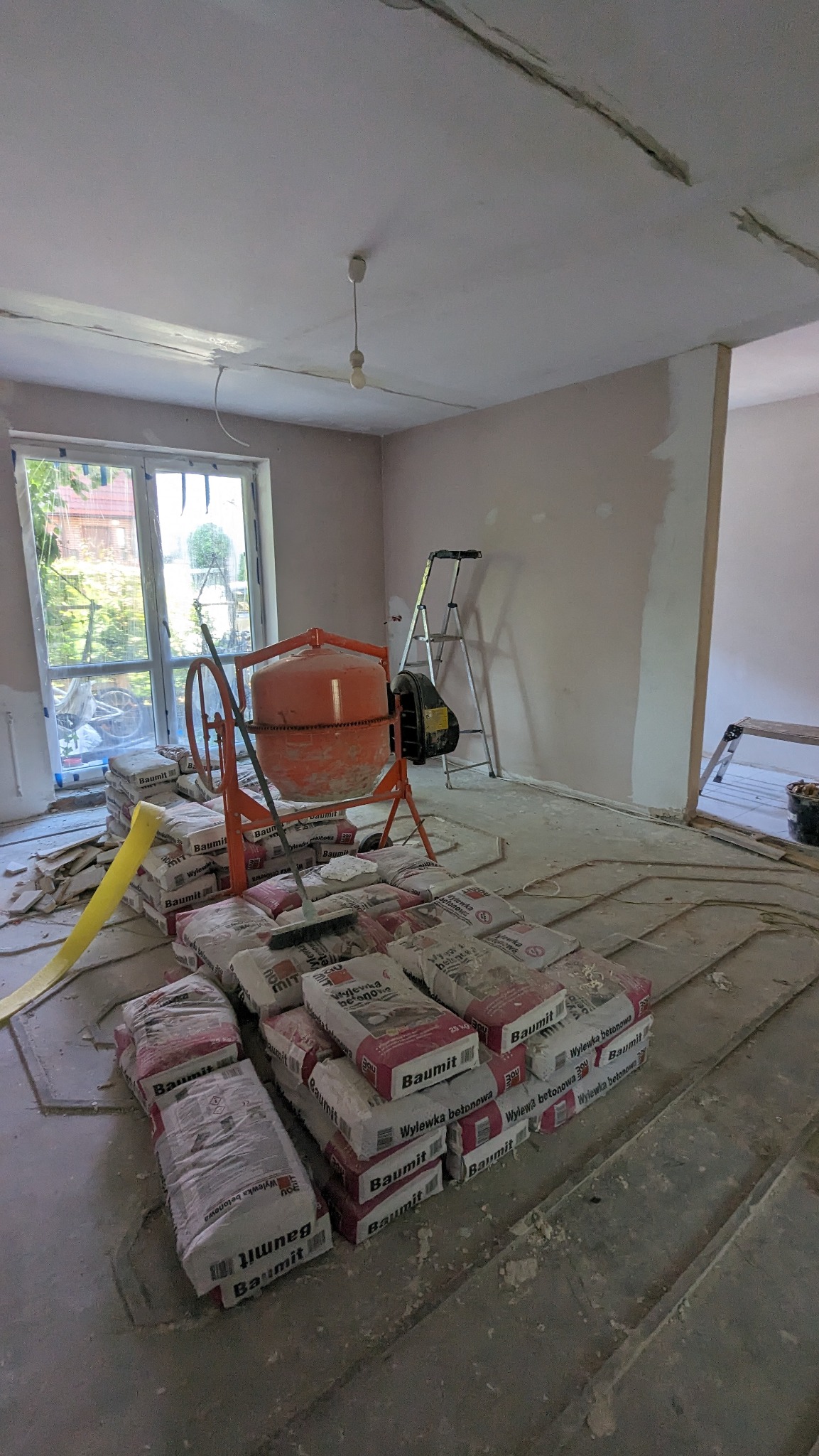 Wnętrze w trakcie wylewki betonowej: worki z zaprawą Baumit, pomarańczowa betoniarka i drabina oparta o ścianę. Widok na okno z zielenią na zewnątrz. Etap prac wykończeniowych.