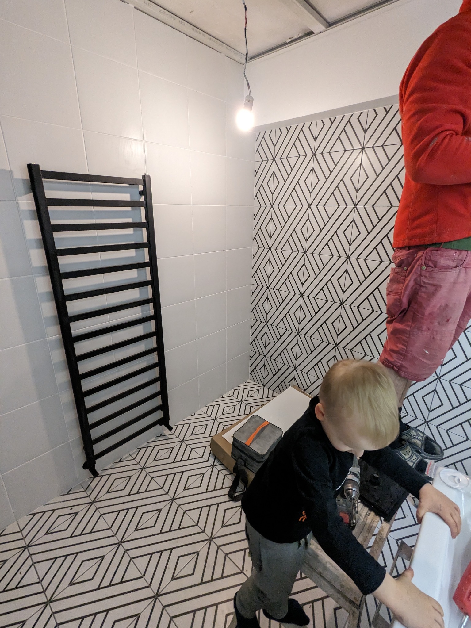 Czarny grzejnik łazienkowy na tle białych płytek i geometrycznego wzoru. Dziecko przygląda się pracy dorosłego na drabinie w trakcie montażu umywalki.