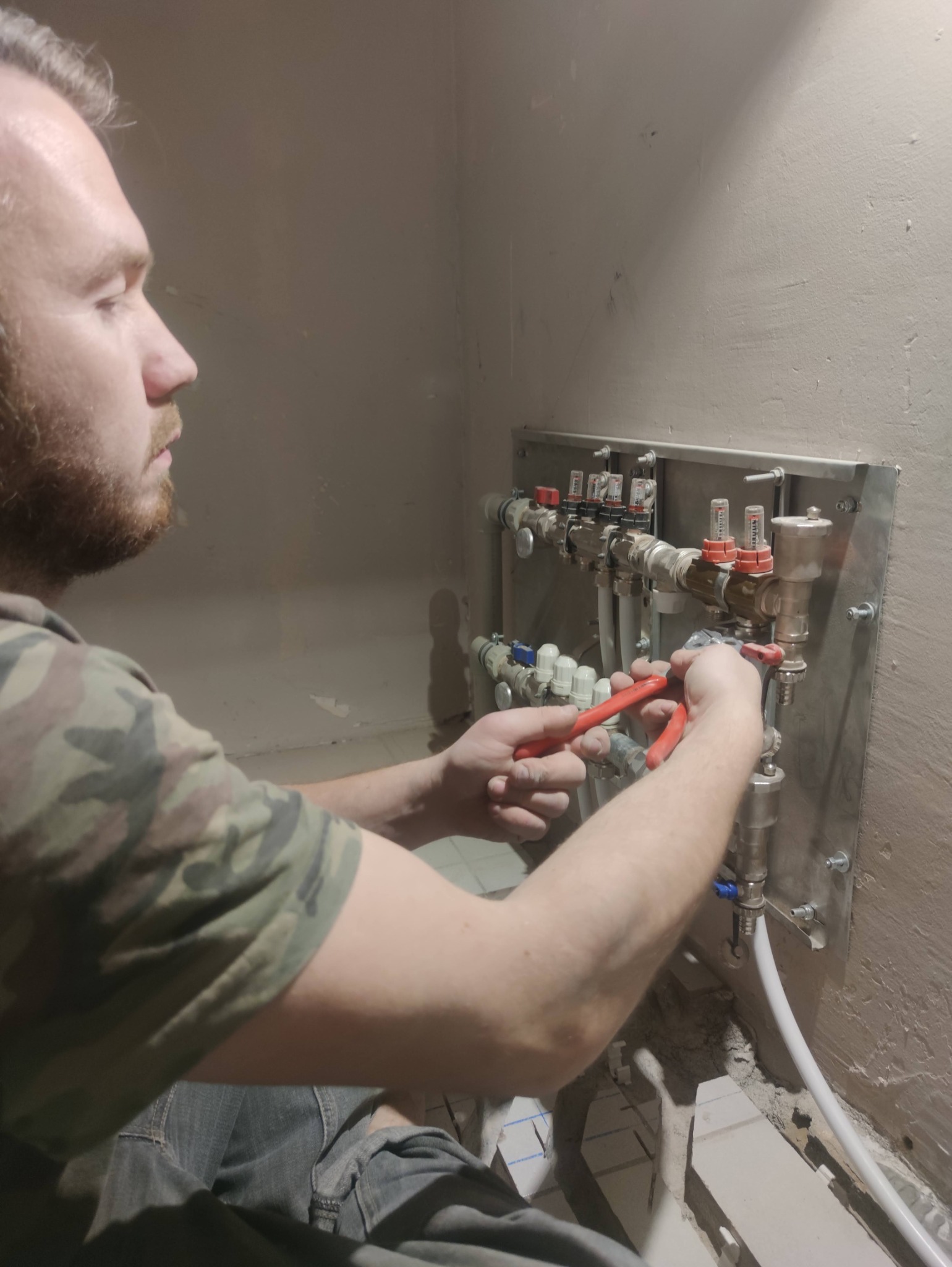 Hydraulik z brodą, w koszulce moro, montuje rozdzielacz ogrzewania podłogowego, używając czerwonych szczypiec. Widoczne białe rury i surowa ściana.