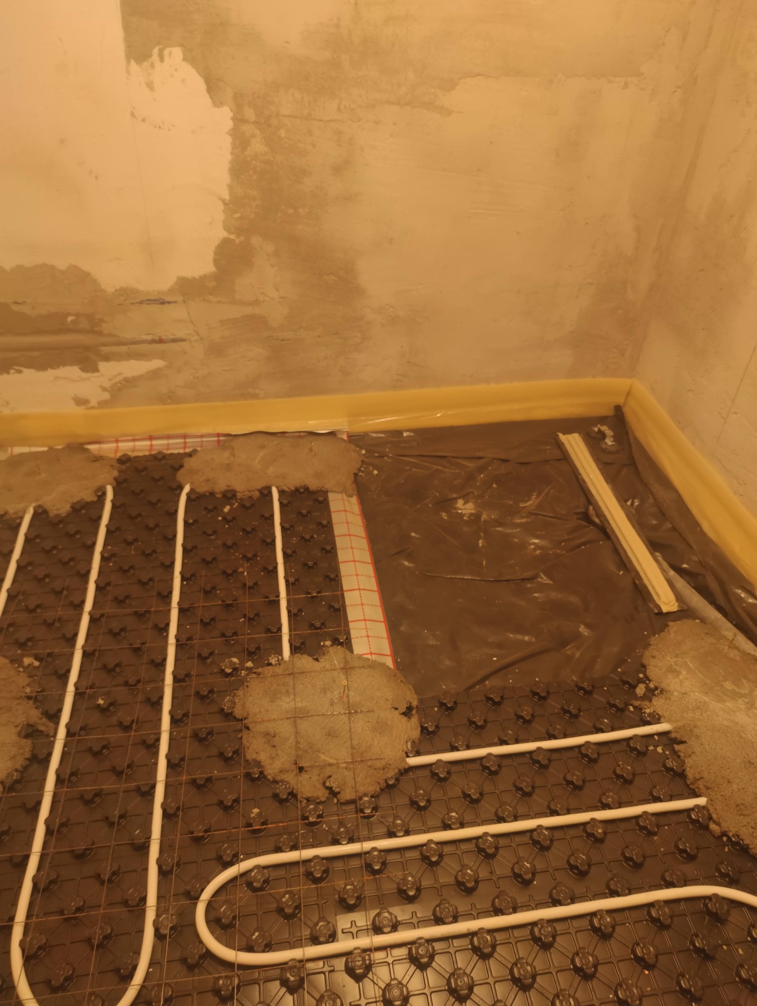Fragment instalacji ogrzewania podłogowego z rurami na czarnej macie, częściowo zalany betonem, widoczne zabezpieczenia folią przy ścianie.