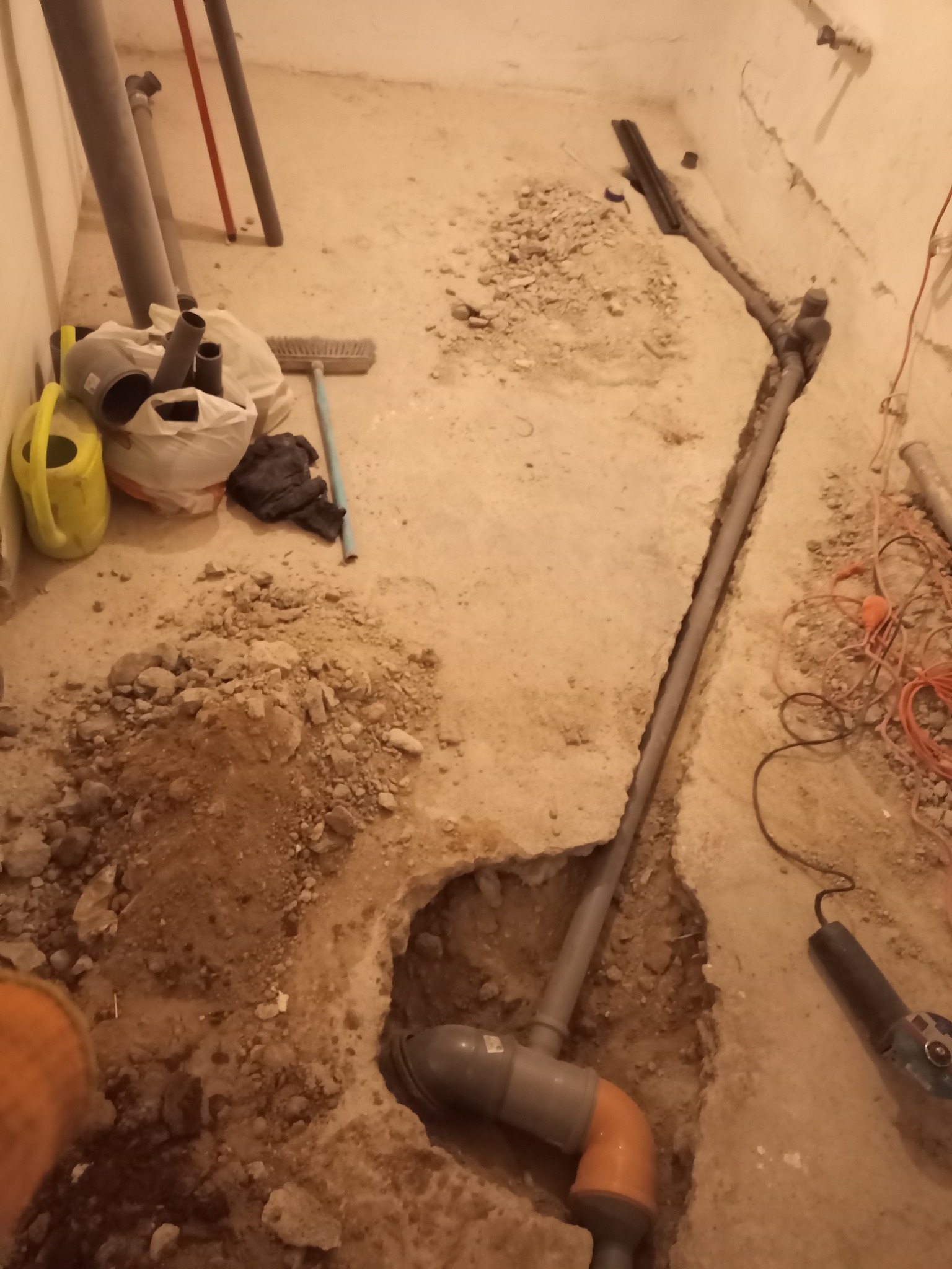 Instalacja rur kanalizacyjnych w wykopie w podłodze. Widoczne rury PCV, kształtki, narzędzia i gruz. Etap prac przed zalaniem posadzki.