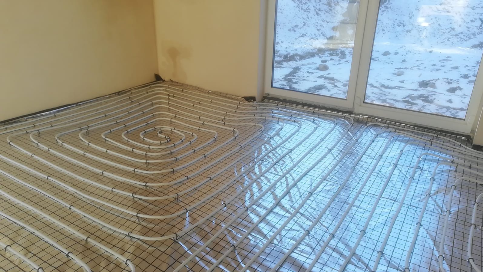 Białe rury ogrzewania podłogowego ułożone spiralnie na siatce, widok z góry, okno z widokiem na zaśnieżony krajobraz w tle, jasne ściany.