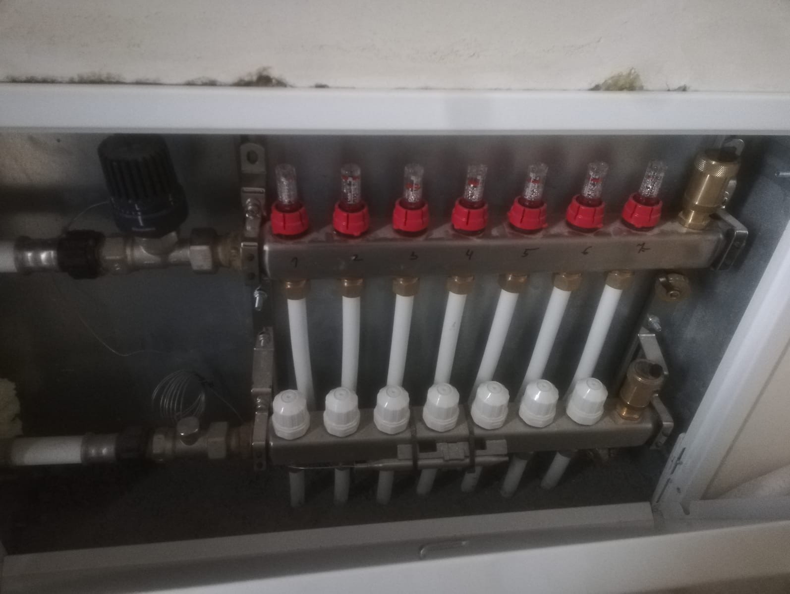 Rozdzielacz ogrzewania podłogowego z rurami w szafce, widok z góry. Czerwone elementy regulacyjne. Metalowa obudowa, białe rury.