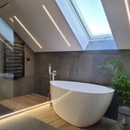 Nowoczesna łazienka na poddaszu z wolnostojącą białą wanną, szarą okładziną ścienną imitującą kamień, oświetleniem LED wzdłuż sufitu i podłogi, kabiną prysznicową ze szklaną ścianą oraz rośliną...