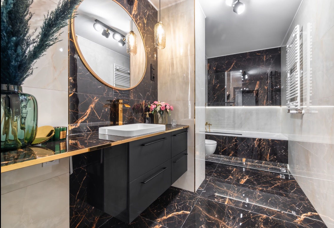 Elegancka łazienka z czarnymi płytkami imitującymi marmur, złotymi akcentami i czarną szafką pod umywalką, oświetlona wiszącymi lampami.